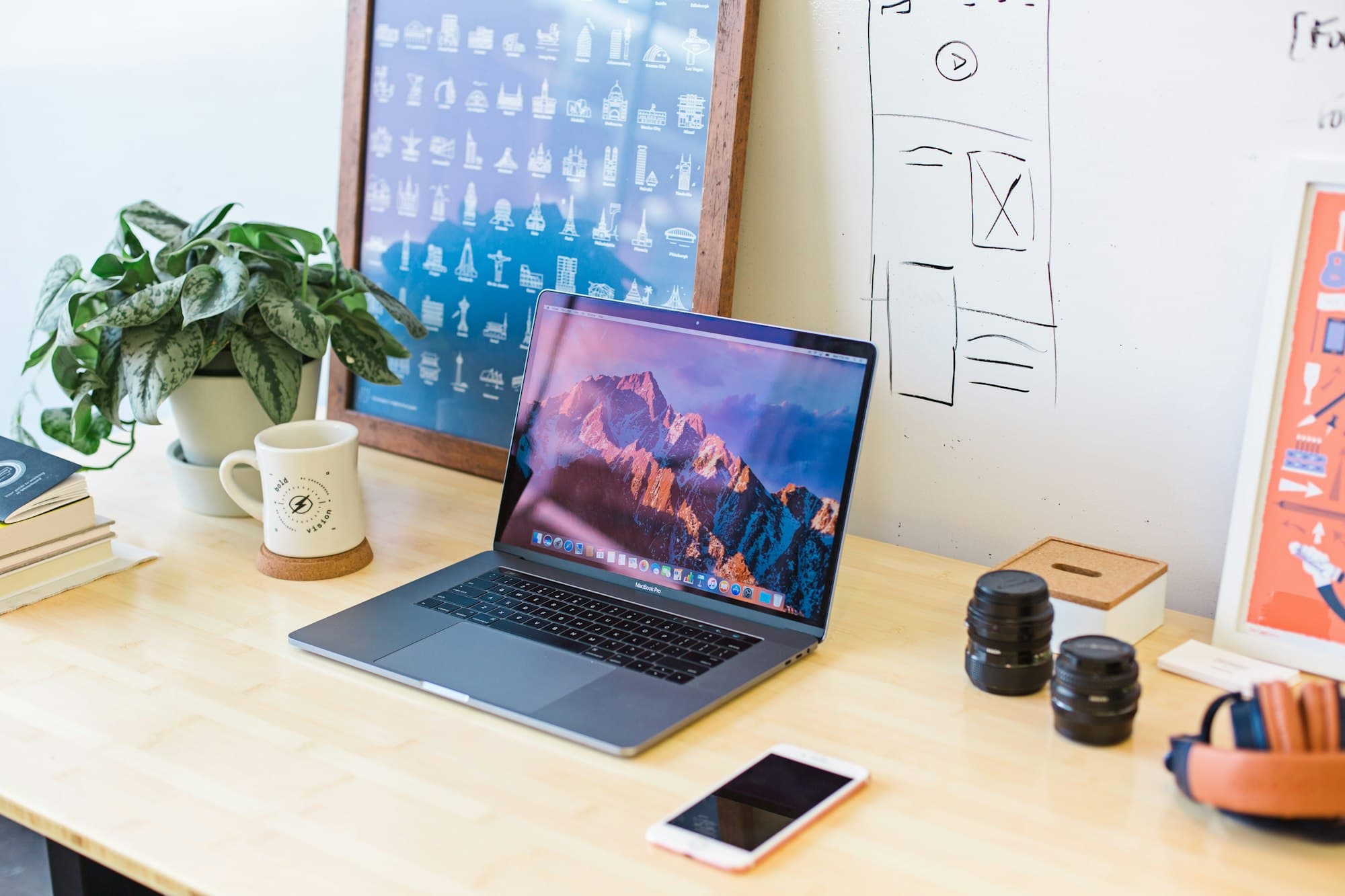 An open laptop on a pale wood desk, beside a plant, a mug and a stack of books