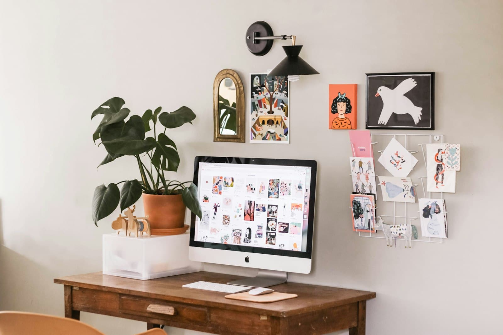 A wooden desk with a computer and a monstera in a terracotta pot, framed prints and postcards pinned to the wall above it